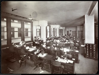 Homens e uma mulher trabalhando em mesas em um escritório da Metropolitan Life Insurance Co. na 23rd Street e Madison Avenue, Nova York, 1907 (impressão em gelatina de prata)